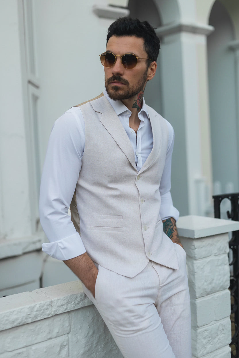 Man wearing a light gray vest over a white shirt and pants, standing against a light-colored building.