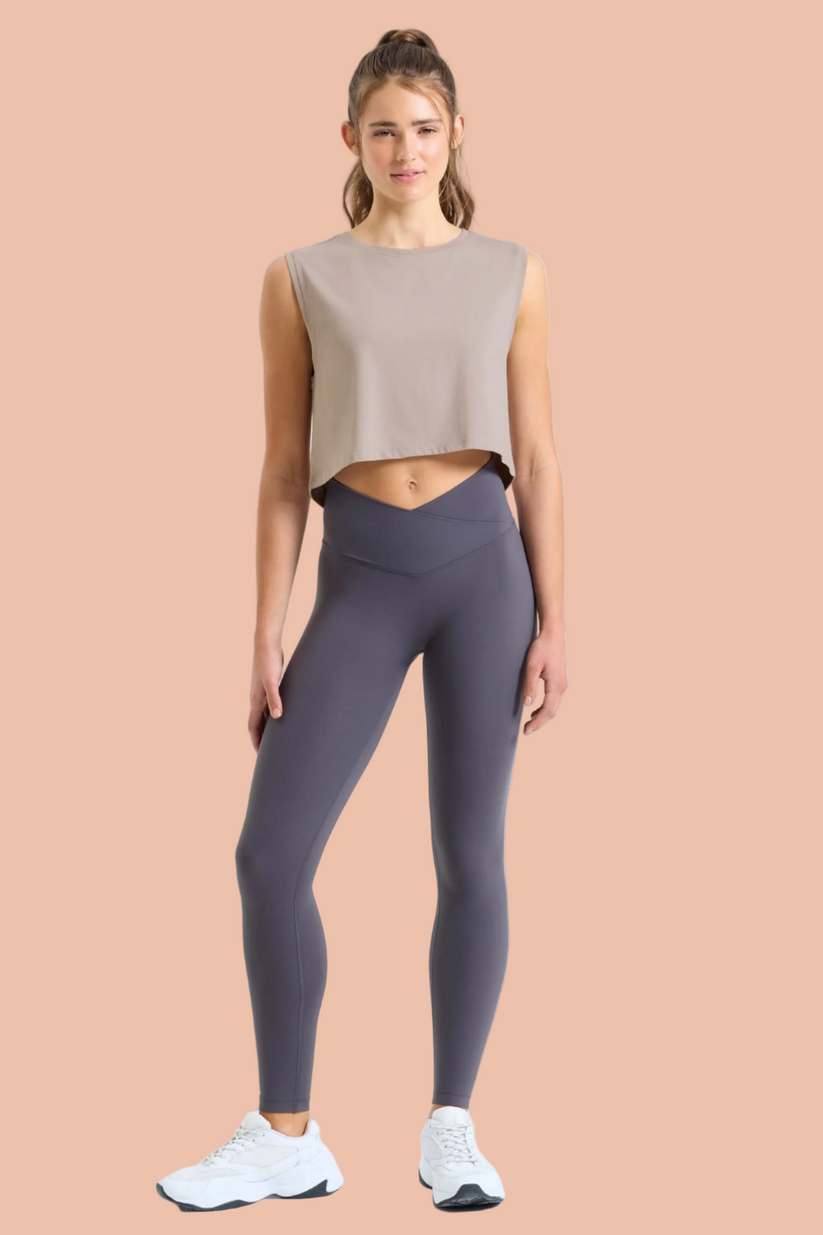 Woman wearing a beige sleeveless top and dark gray leggings on a beige background