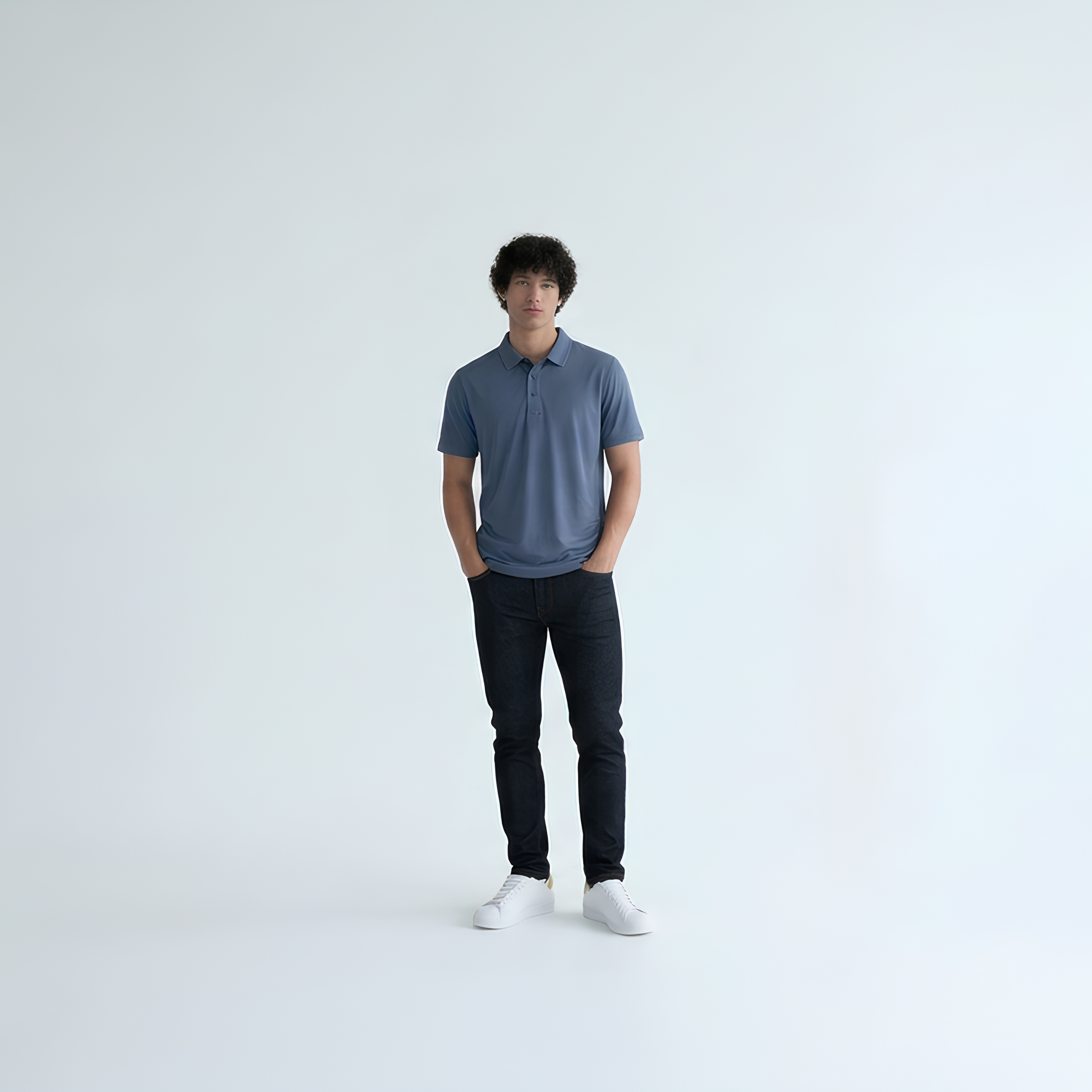 A man wearing a slate blue short-sleeve polo shirt, black pants, and white sneakers standing against a seamless white studio background.