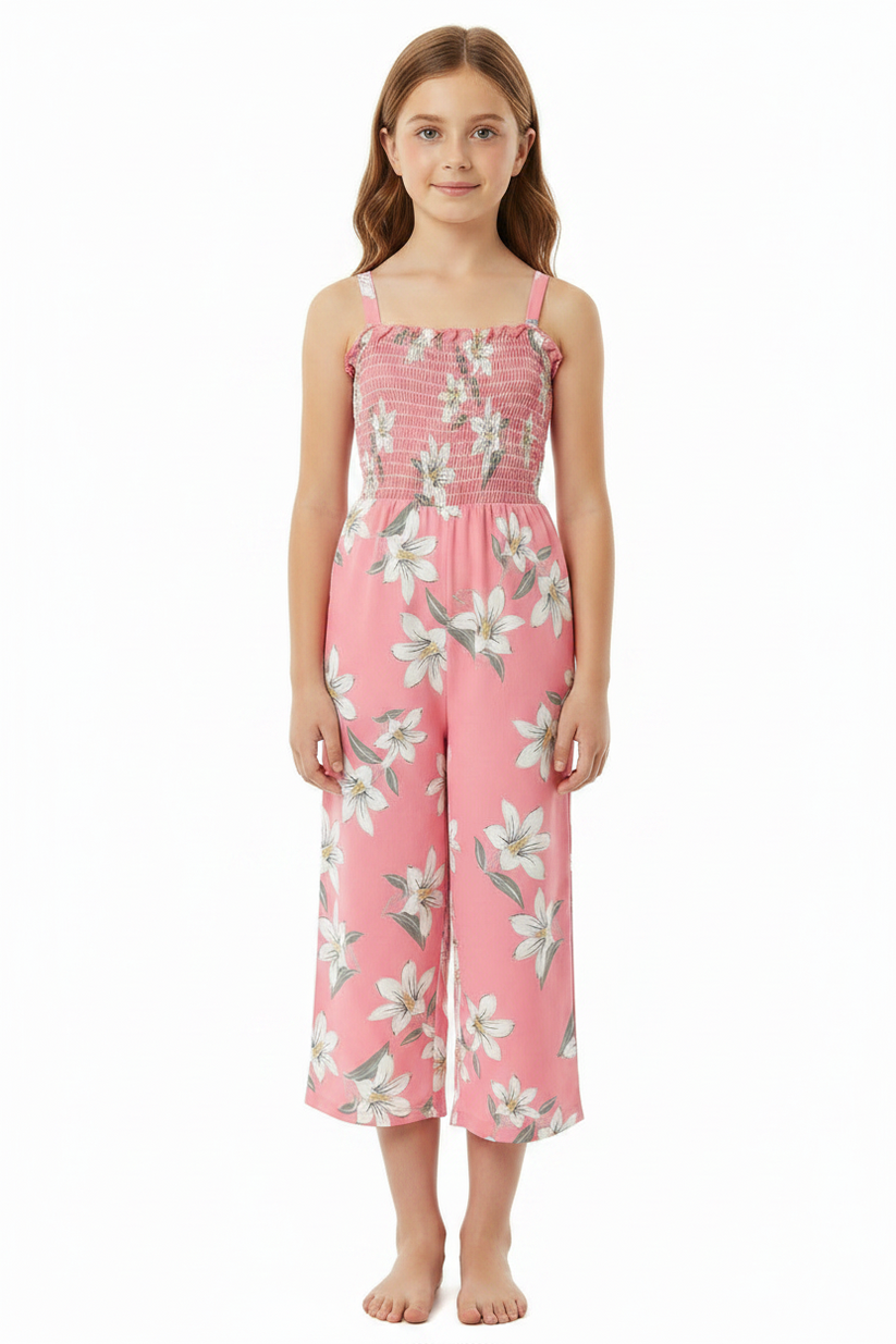 A young girl modeling a pink floral jumpsuit with white flowers and green leaves, featuring a smocked bodice and wide legs, set against a plain white background.