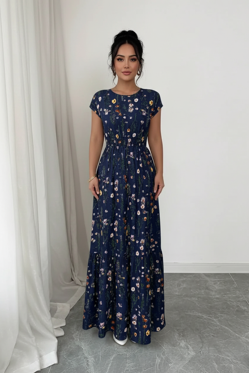 A woman modeling a navy blue maxi dress with a white and multicolor floral pattern, standing in a minimalist room with white walls and a gray floor.