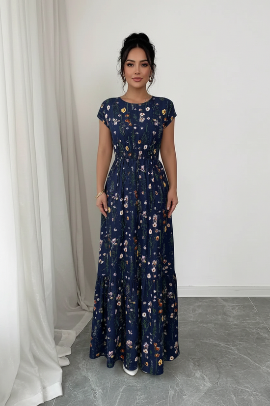A woman modeling a navy blue maxi dress with a white and multicolor floral pattern, standing in a minimalist room with white walls and a gray floor.