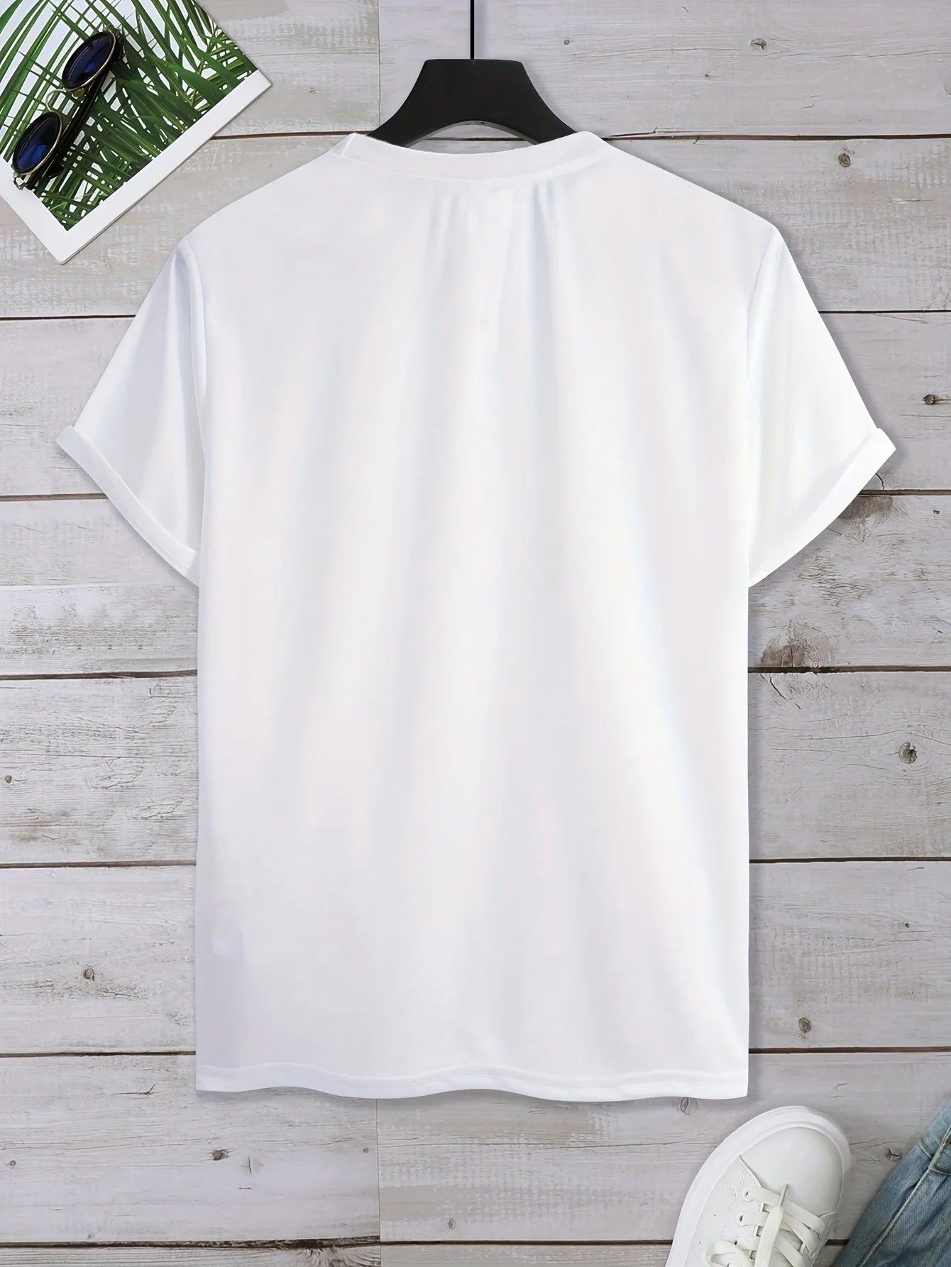 A plain white short-sleeve t-shirt hanging on a black hanger against a light gray wood plank background, with partial styling props visible at the corners.
