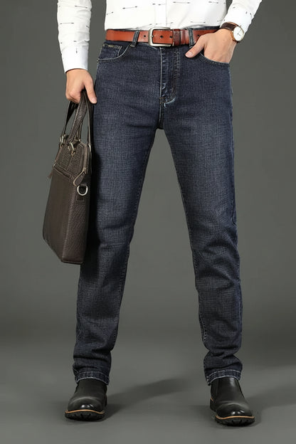 Person wearing blue jeans and a white shirt with a brown leather belt, holding a brown leather bag on a gray background.