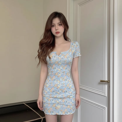 A woman with long wavy light brown hair wearing a light blue floral print mini dress, posing in front of a white paneled door.