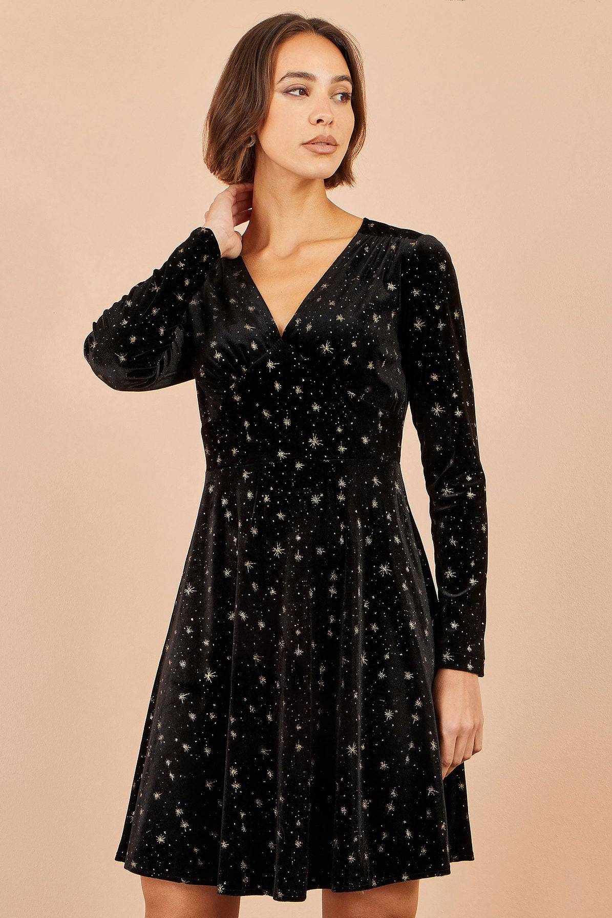 A woman modeling a black velvet long-sleeve V-neck dress with a scattered gold star pattern, set against a solid beige background.