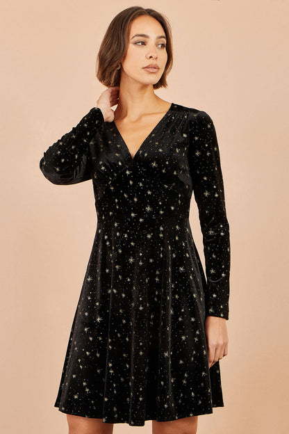 A woman modeling a black velvet long-sleeve V-neck dress with a scattered gold star pattern, set against a solid beige background.