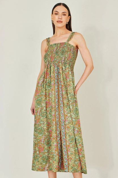 A woman modeling a sleeveless midi dress with a green tropical floral print, featuring a smocked bodice and a straight neckline, set against a plain light gray background.