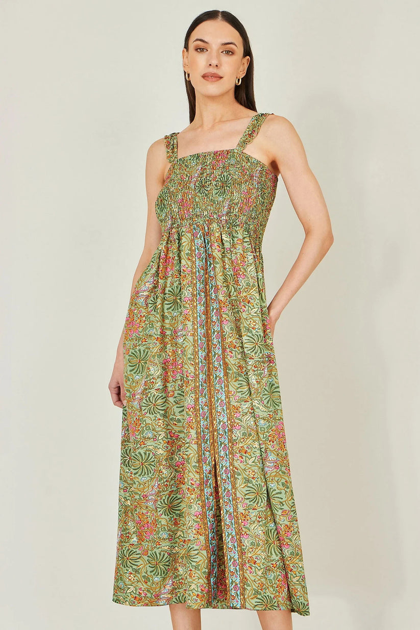 A woman modeling a sleeveless midi dress with a green tropical floral print, featuring a smocked bodice and a straight neckline, set against a plain light gray background.